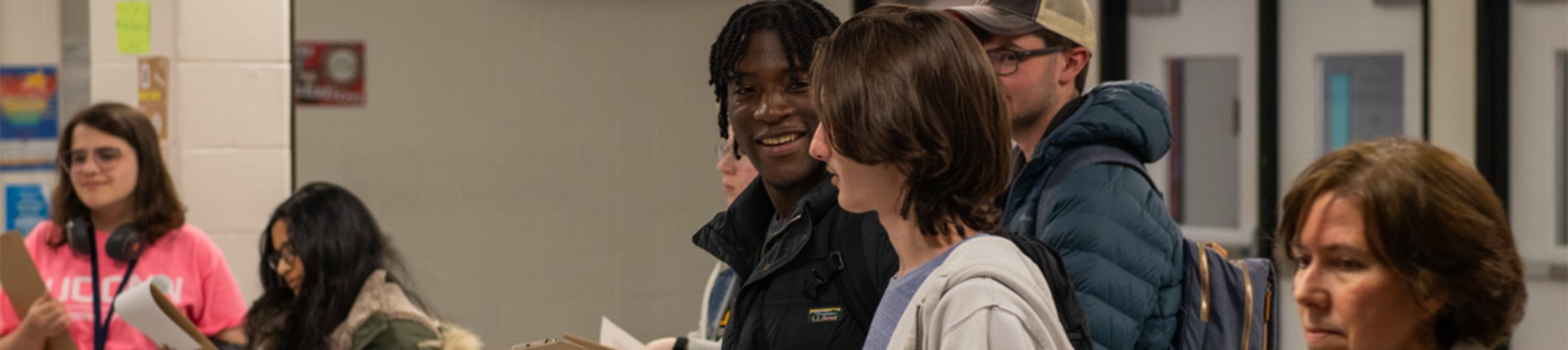 Male high school students stand together, talking and smiling. A woman and two female college students stand next to them; the college students hold clipboards.