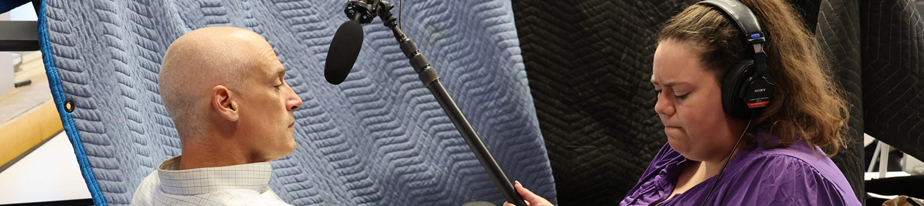 A woman holds a microphone in front of a man while holding an audio recorder.